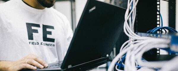 Un ingénieur en T-shirt blanc marqué 'FE' utilise un ordinateur portable devant des serveurs avec des câbles bleus.
